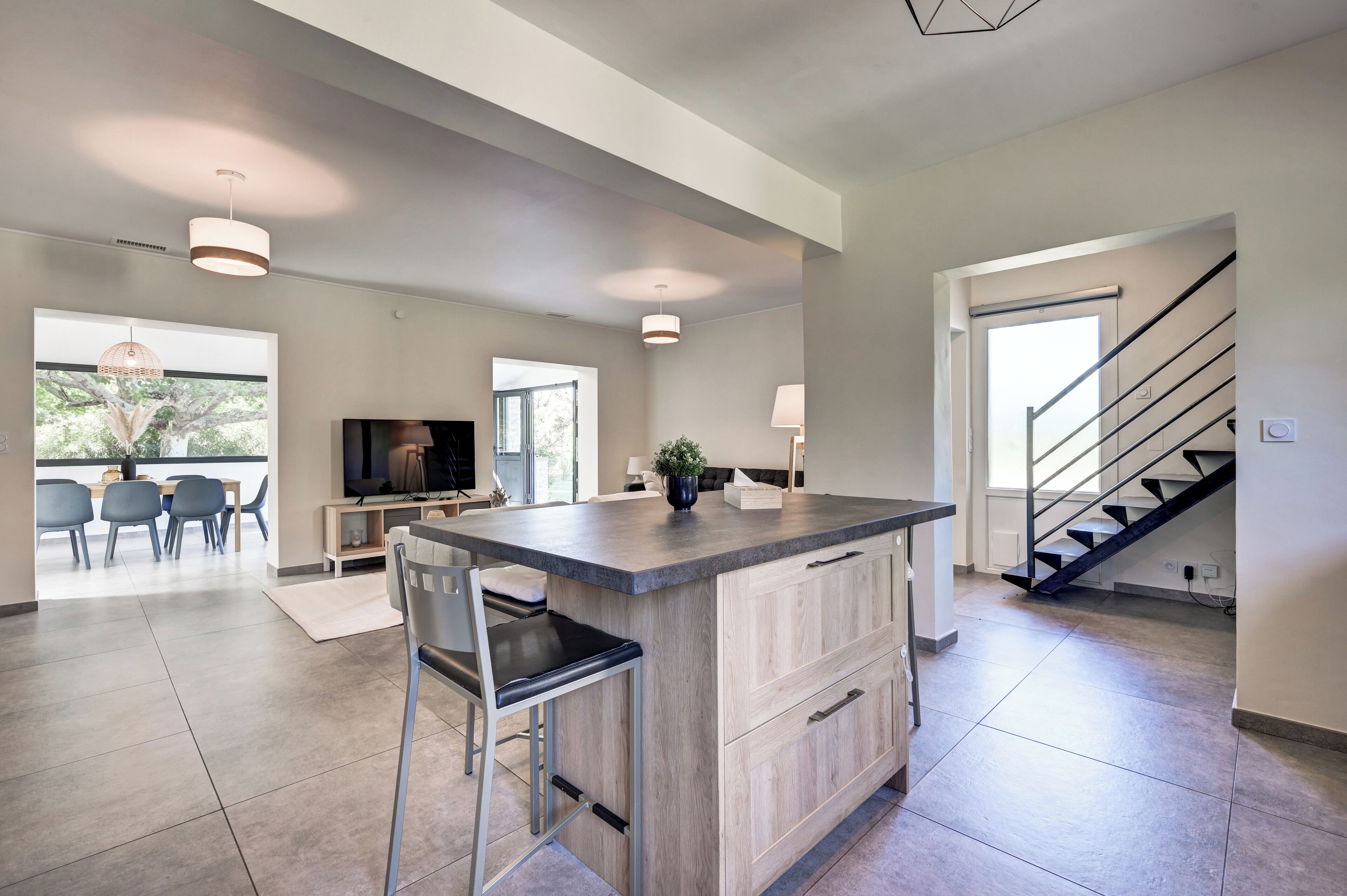 Una cocina moderna con una isla central, un comedor con sillas, un televisor y una escalera que conduce a otro nivel.