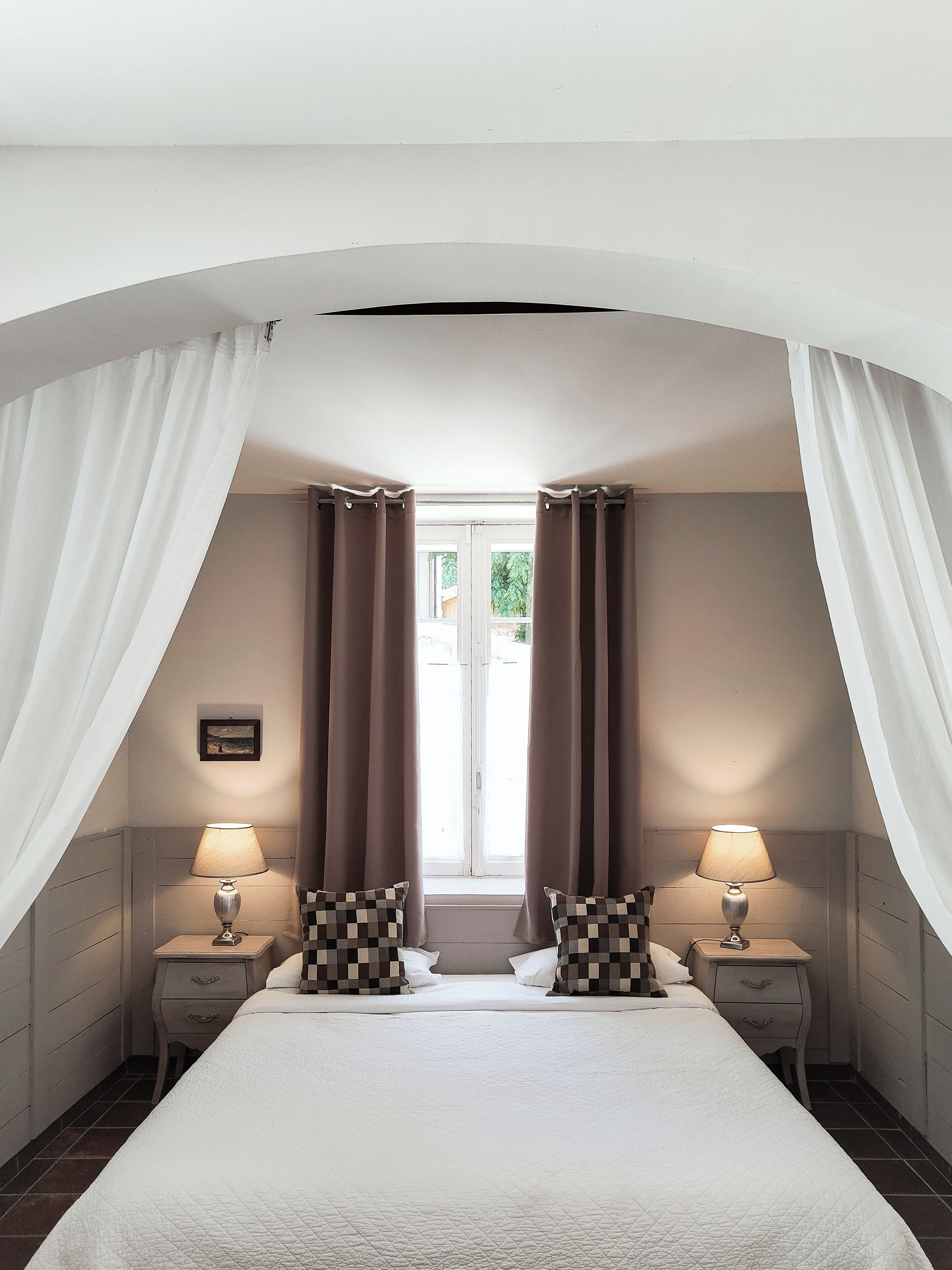 A bedroom with a large bed, two bedside tables with lamps, a window with curtains, and a picture on the wall.