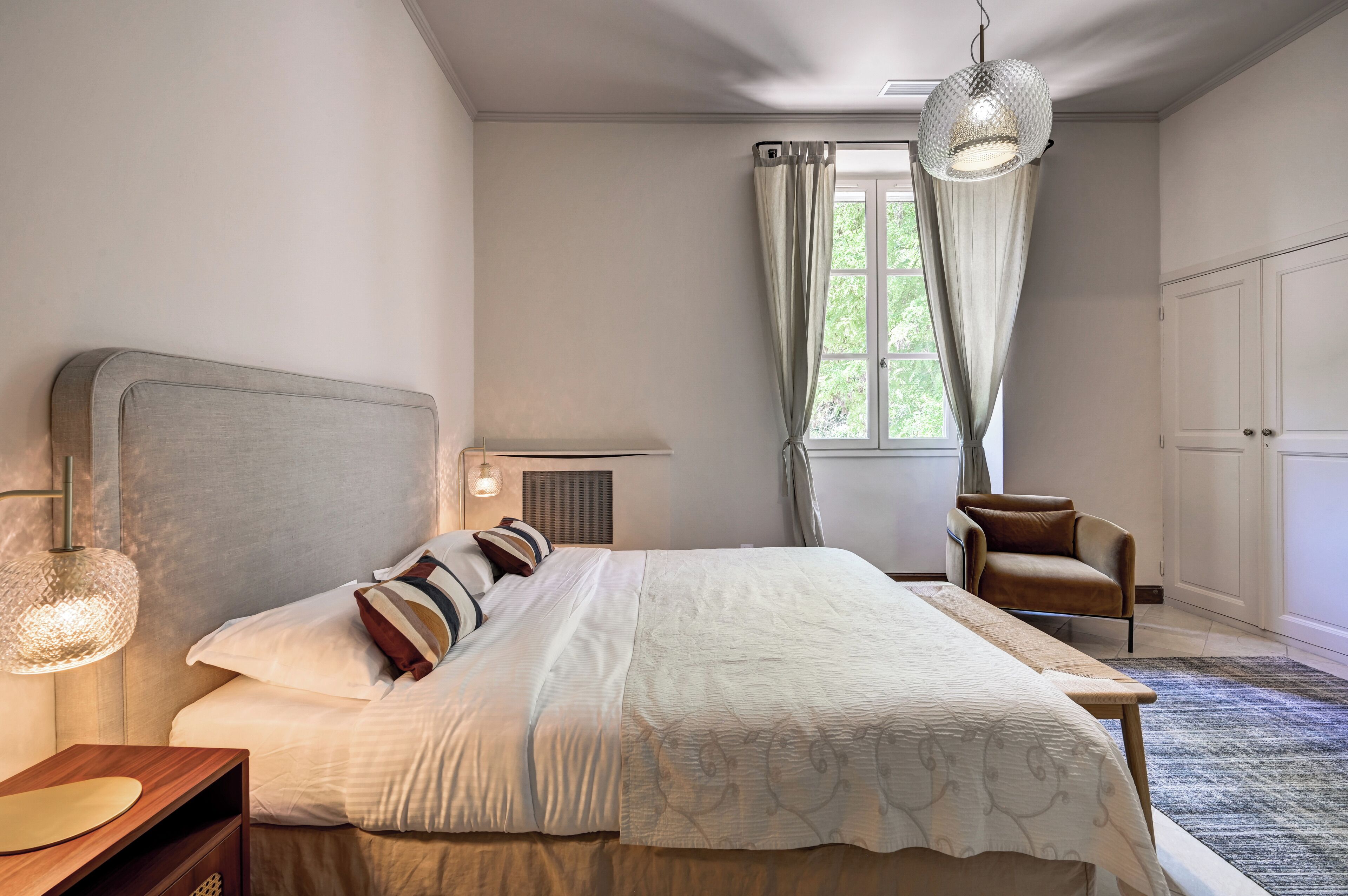 Um quarto com uma cama grande, uma mesa de cabeceira, uma cadeira, um lustre e uma janela com cortinas.