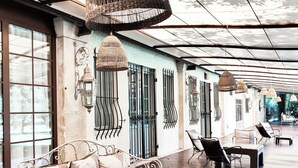 A covered outdoor seating area with a wicker hanging light fixture, metal-framed furniture, and a view of the street through large windows. - Mas des Comtes de Provence  (Tarascon)