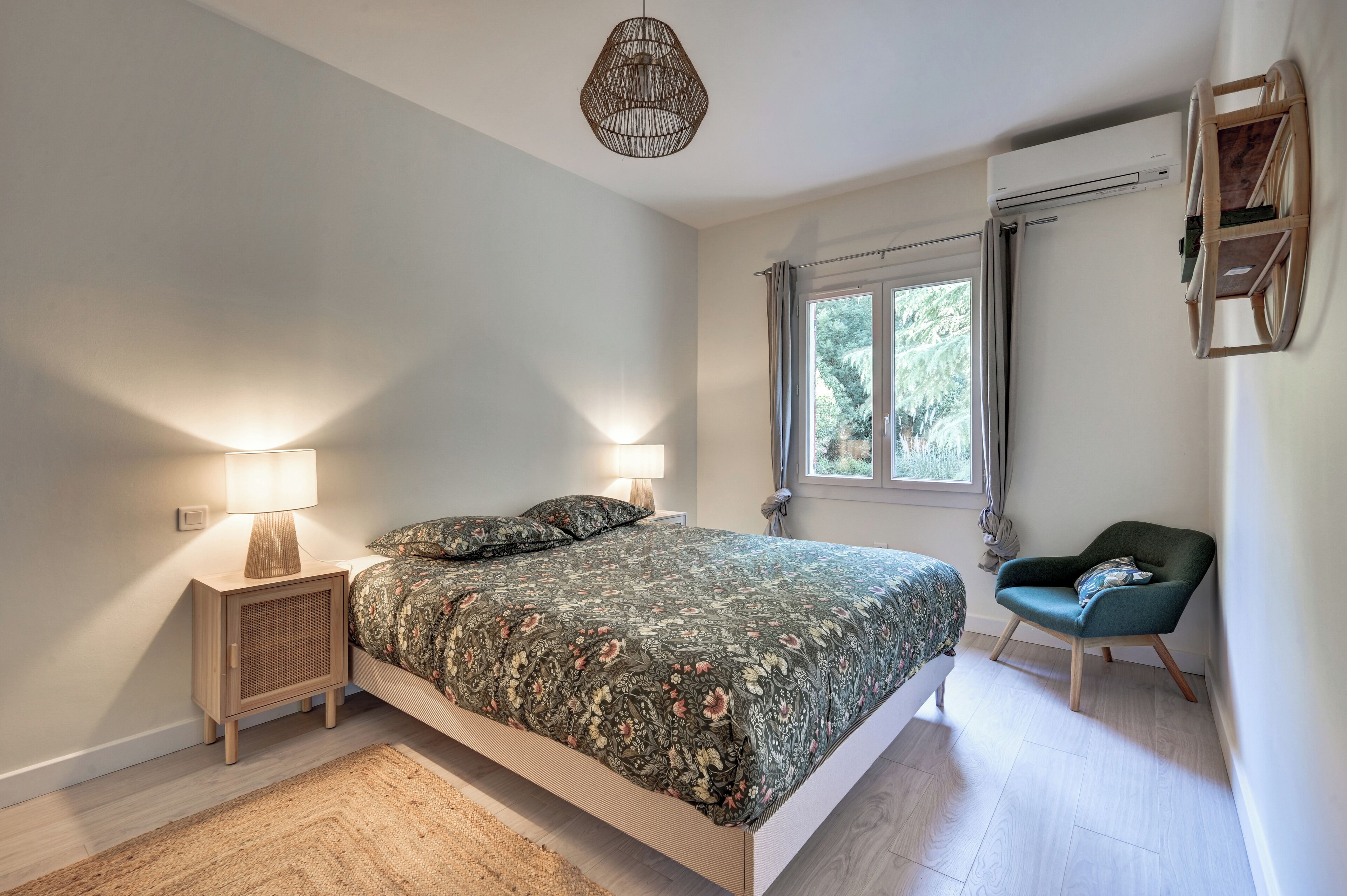 A bedroom with a bed, bedside lamps, a chair, a window, and an air conditioning unit.