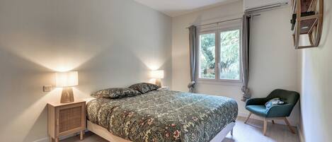 A bedroom with a bed, bedside lamps, a chair, a window, and an air conditioning unit.