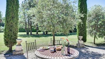 Een ronde tafel met een tafelkleed met patroon, omringd door een laag metalen hekwerk, gelegen in een tuin met hoge bomen en een grindpad.