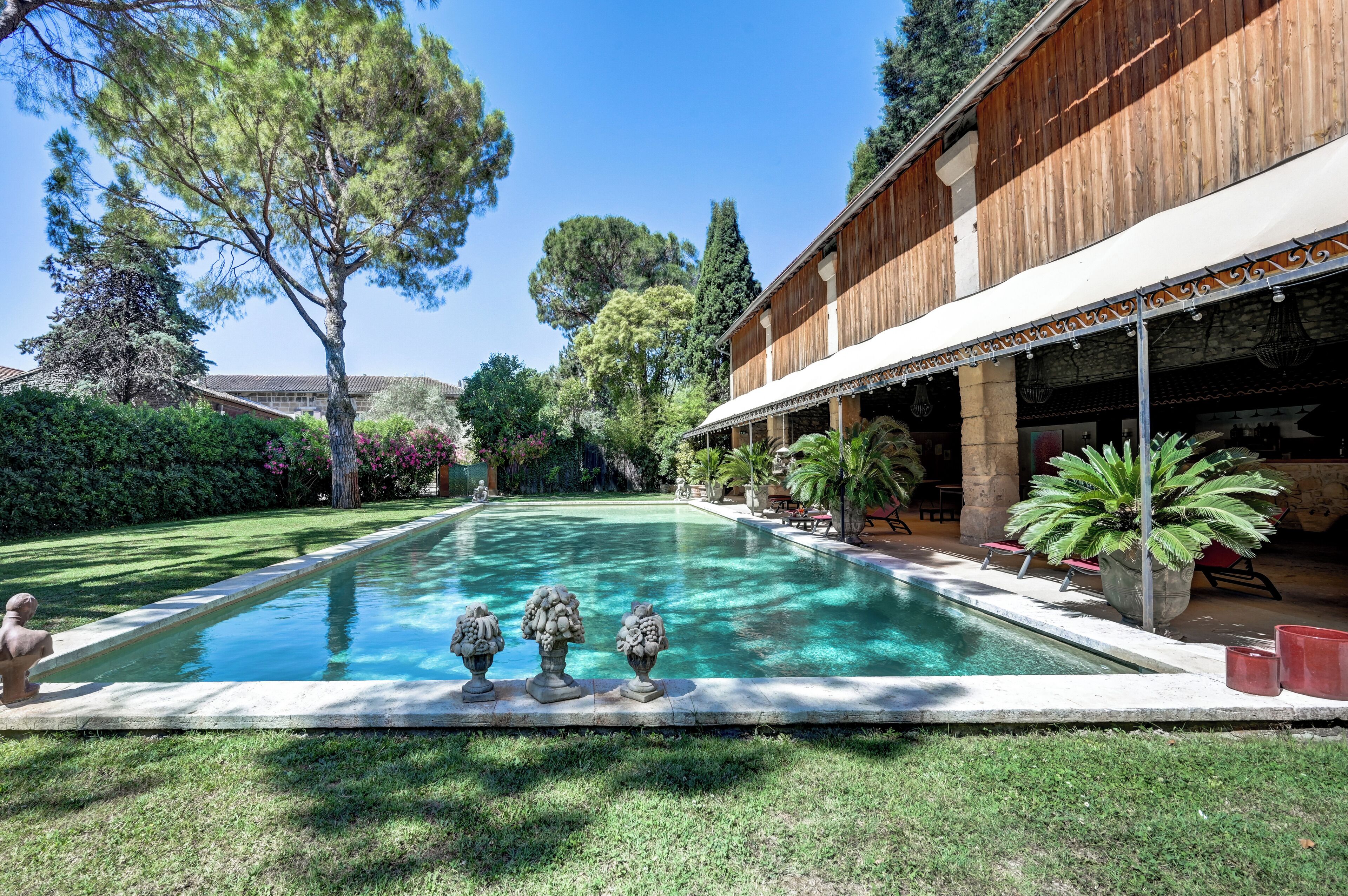 A swimming pool with decorative urns, a wooden building with a covered patio, and lush greenery.