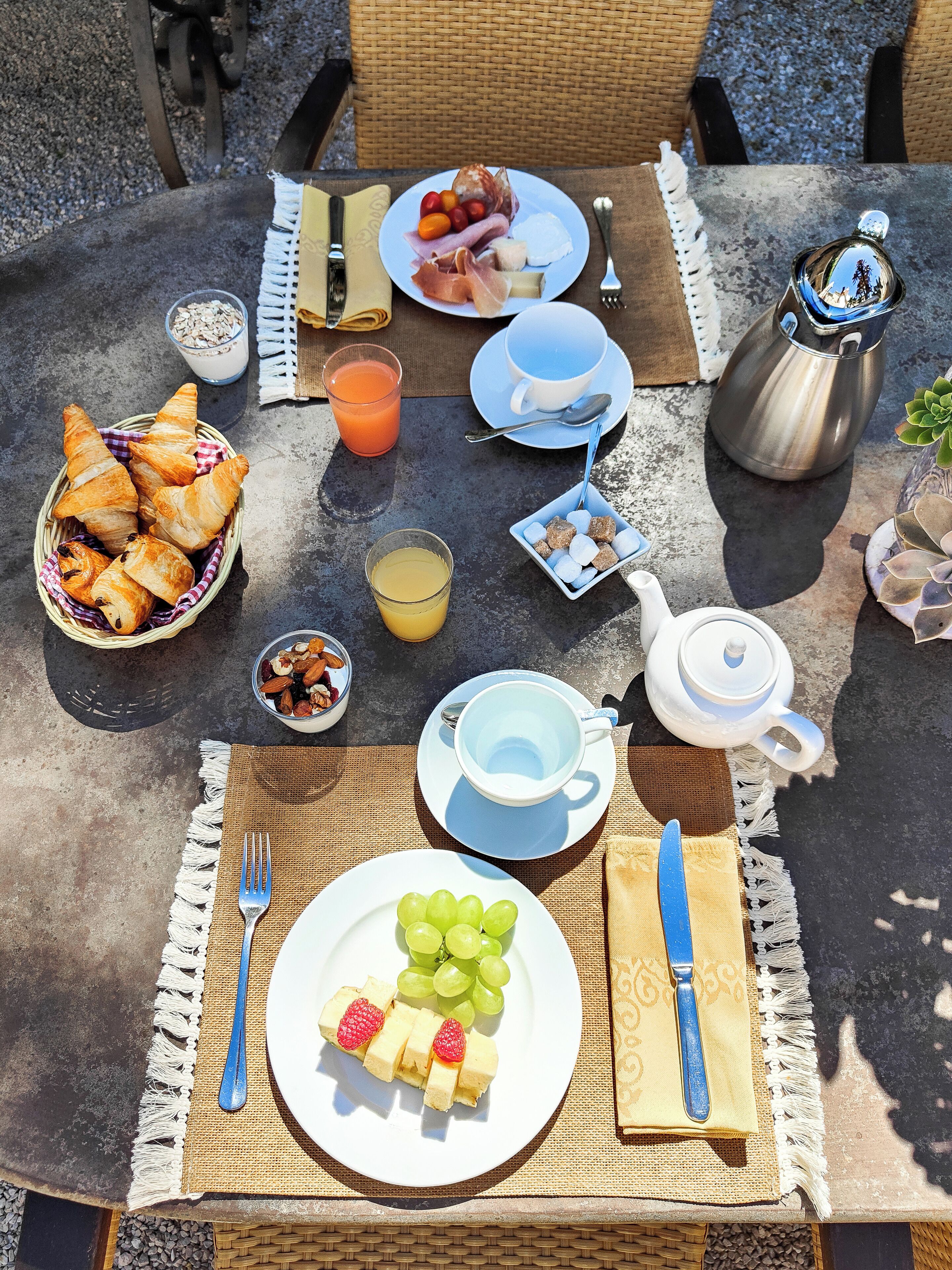 Une table dressée à l’extérieur, avec des aliments pour le petit-déjeuner, notamment des croissants, des fruits et une théière.