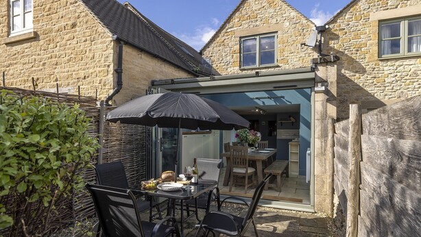 Une maison en pierre avec une porte en verre donnant sur un coin repas équipé d’une table, de chaises et d’un parasol.