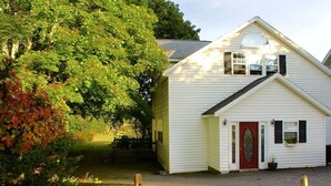 A white two-story house with a red door, a small porch, and a garden area with trees and shrubs. - Songbrook + (Otego)