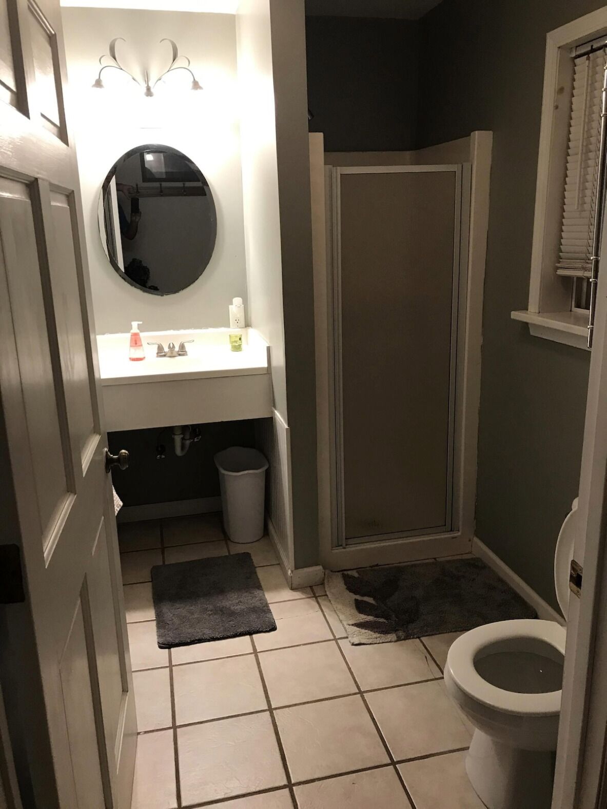 A bathroom with a toilet, a sink with a mirror, a shower, and a trash can.