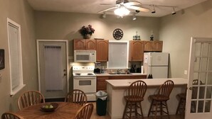 A compact kitchen with wooden cabinets, a microwave, a stove, and a refrigerator. There is a dining table with chairs and a small island with bar stools. - Songbrook + (Otego)