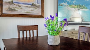 Ett matbord med en vas med lila blommor, två trästolar och en inramad tavla som föreställer en strandscen på väggen.