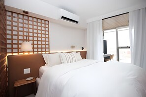 Una habitación de hotel con una cama grande, una mesita de noche, aire acondicionado de pared, un panel de madera con diseño de cuadrícula y un ventanal con cortinas.