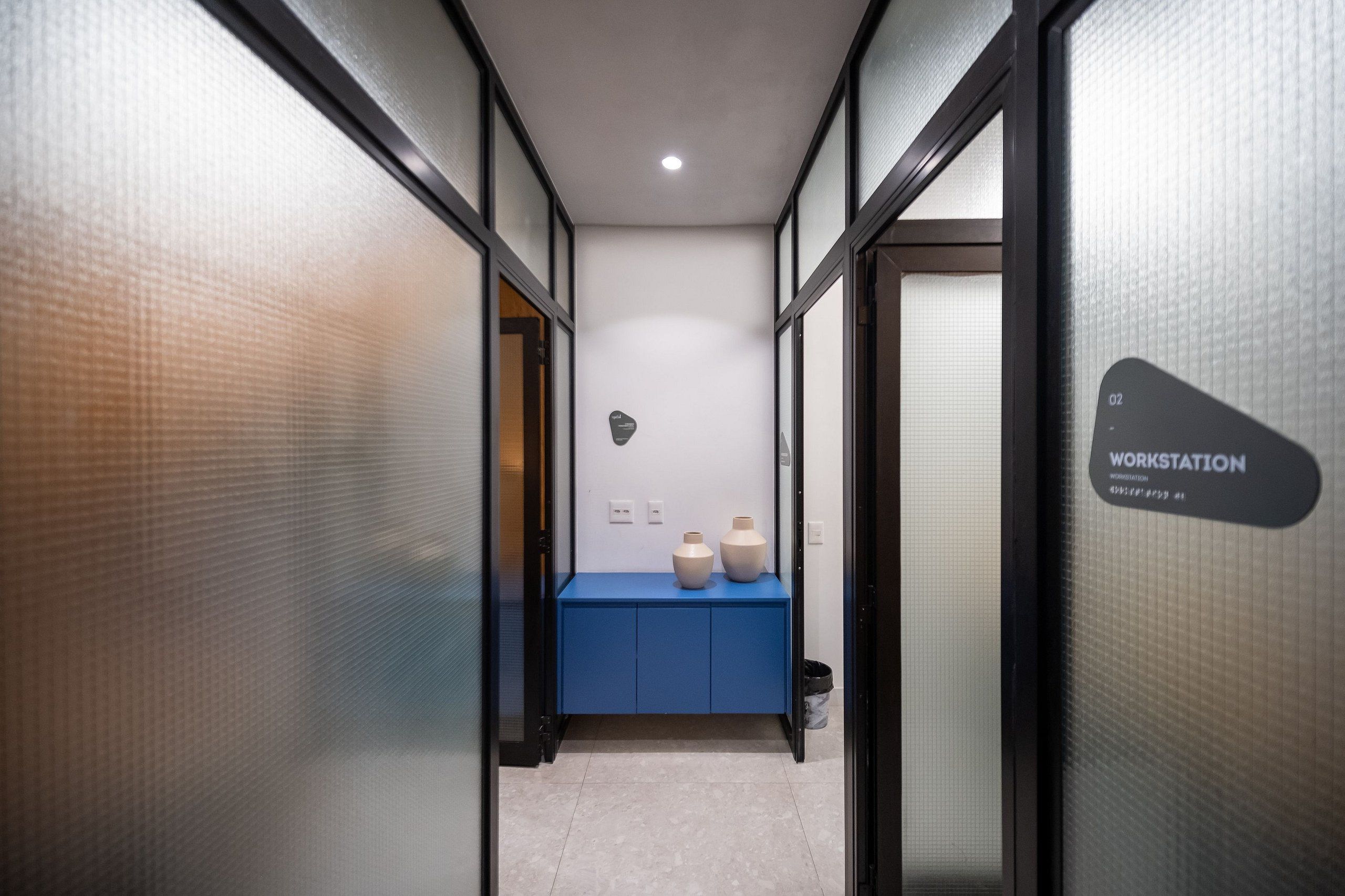 Un couloir avec un comptoir bleu, deux vases en céramique et un panneau indiquant « POSTE DE TRAVAIL ».