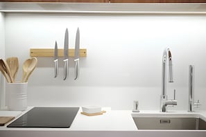 Una cocina moderna con encimera blanca, tabla de cortar negra, utensilera de madera, pileta de acero inoxidable y un grifo.
