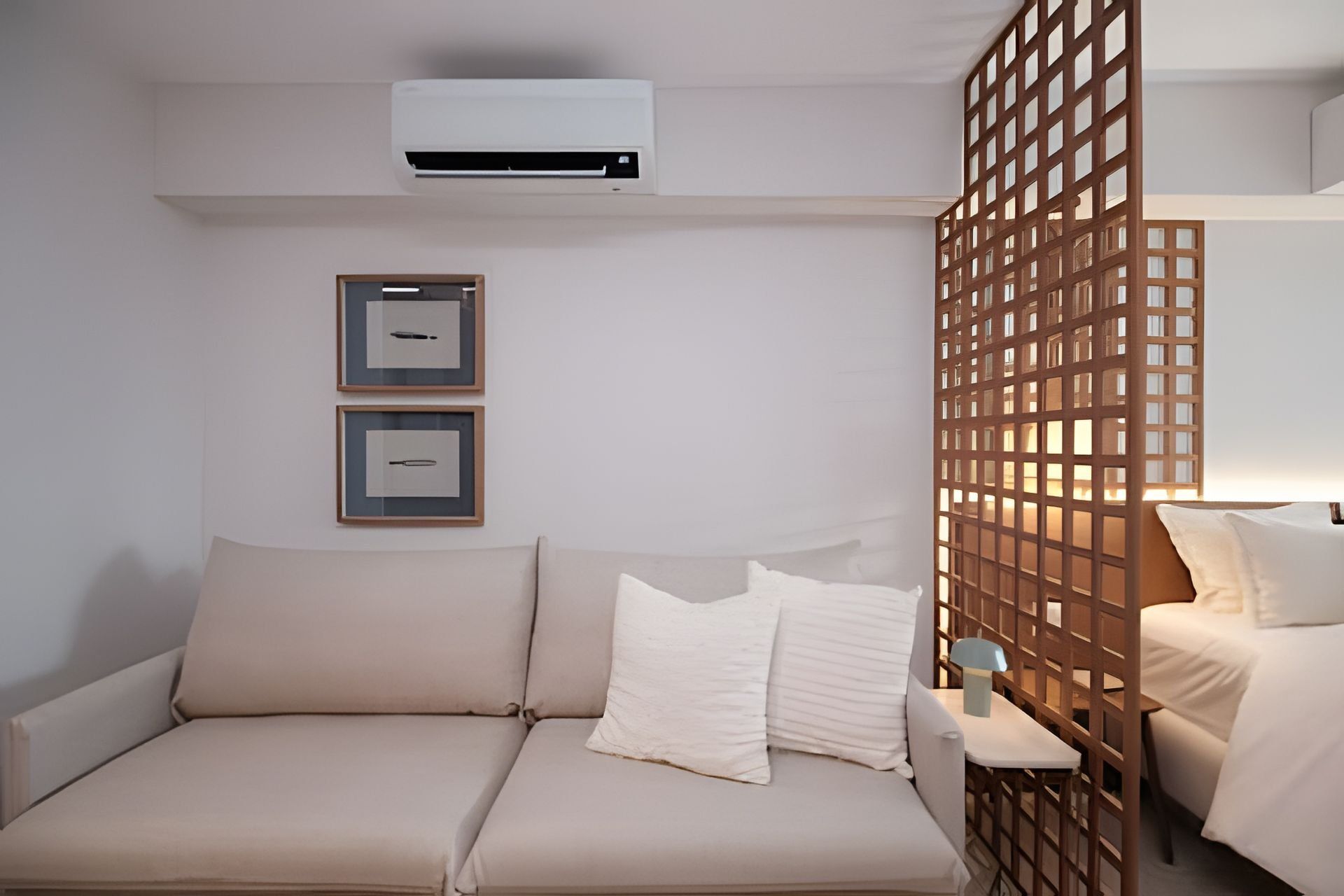 A hotel room with a beige sofa, two framed pictures on the wall, an air conditioning unit, and a wooden partition.