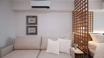 A hotel room with a beige sofa, two framed pictures on the wall, an air conditioning unit, and a wooden partition.