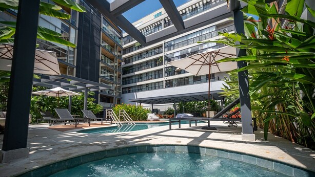 Zona de piscina moderna de hotel con un jacuzzi central, sillones de descanso y un edificio de varios pisos al fondo.