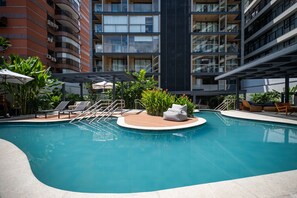 Un área de piscina moderna con sillones de descanso y una pequeña zona de estar, rodeada de edificios altos.