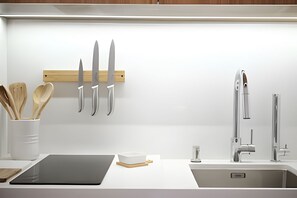 Una cocina moderna con encimera blanca, tabla de cortar negra, utensilera de madera, pileta de acero inoxidable y un grifo.