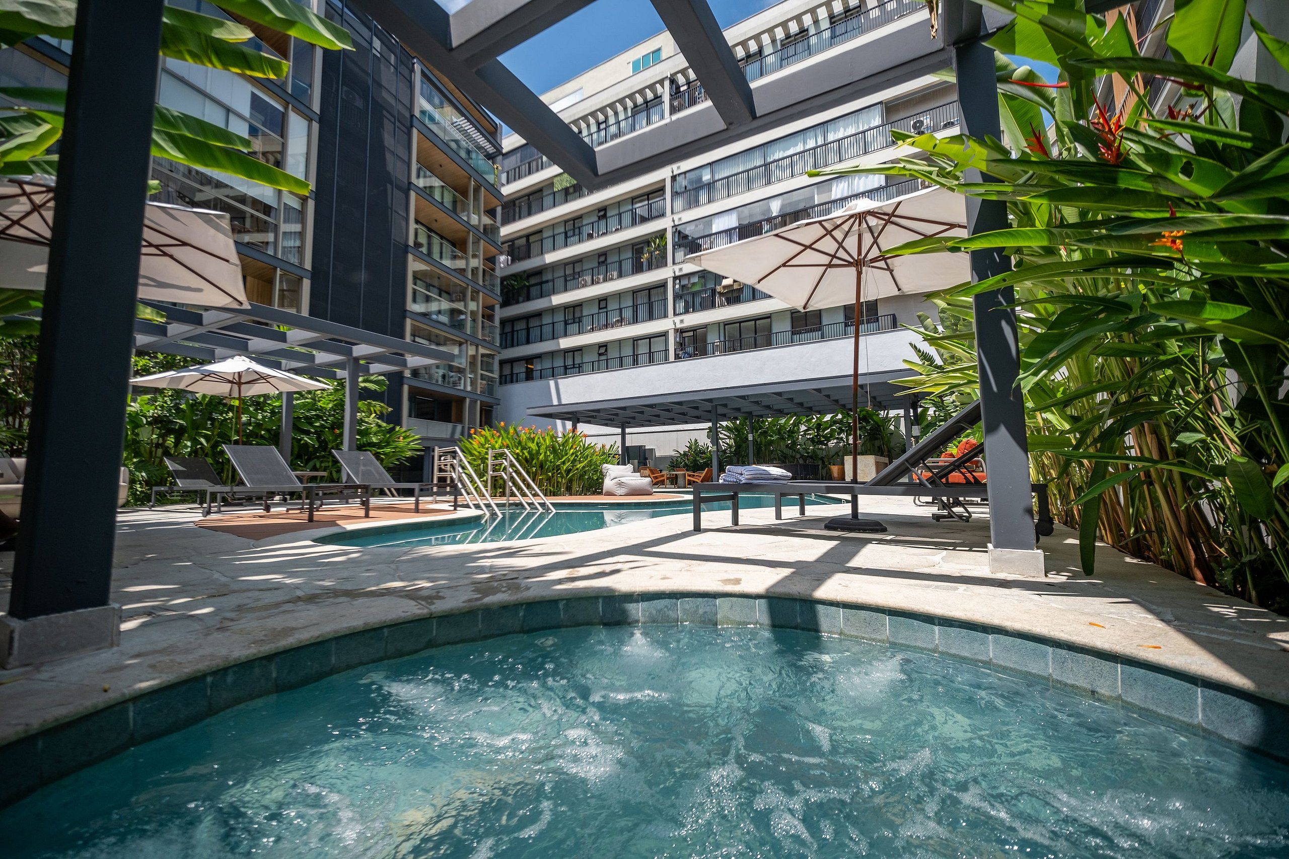 A modern hotel pool area with a central hot tub, lounge chairs, and a multi-story building in the background.