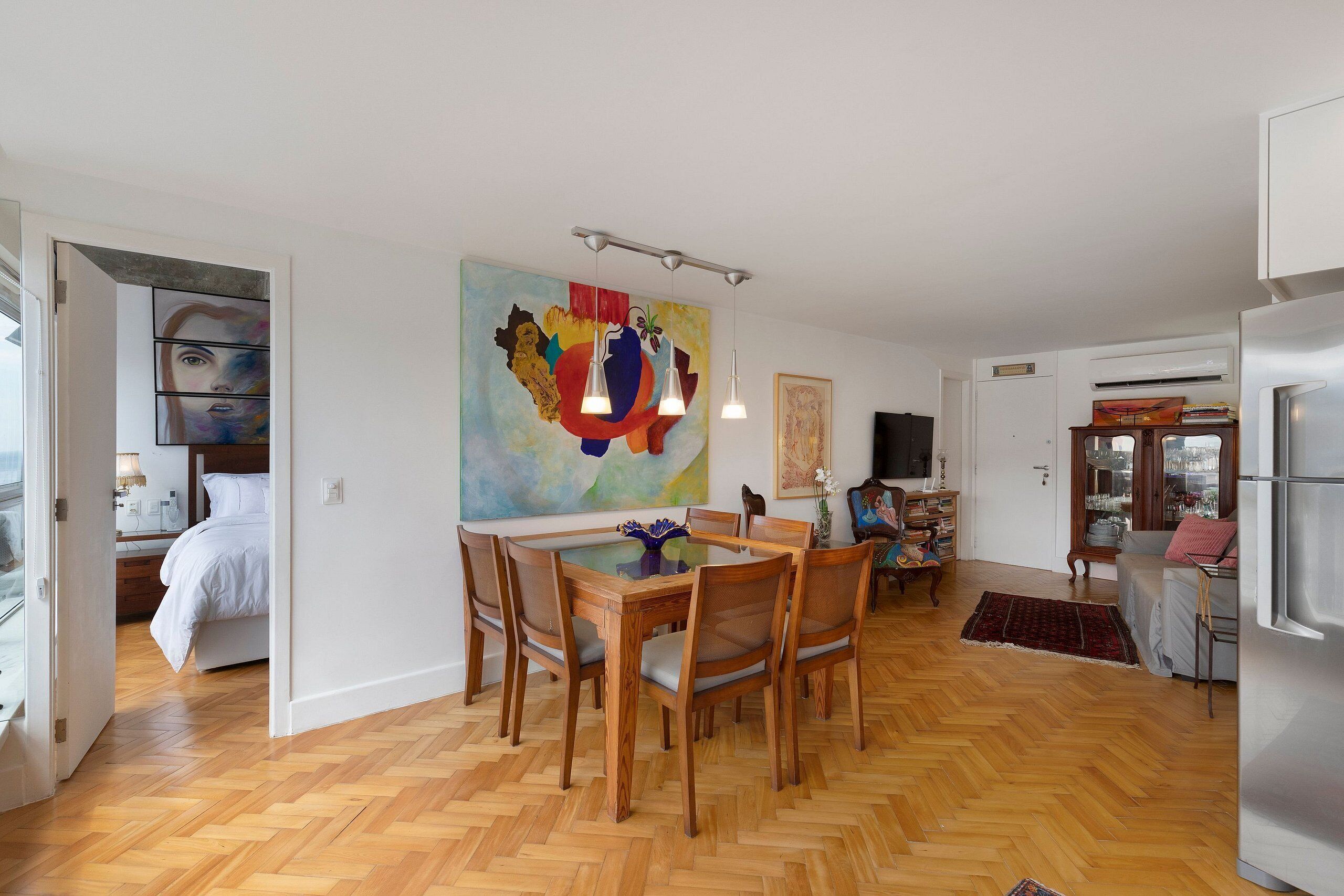 Un amplio salón con suelo de parqué labrado, una mesa de comedor con superficie de vidrio rodeada de sillas de madera y un vibrante cuadro abstracto en la pared.