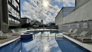 Een zwembad met ligbedden en een jacuzzi, omgeven door gebouwen.