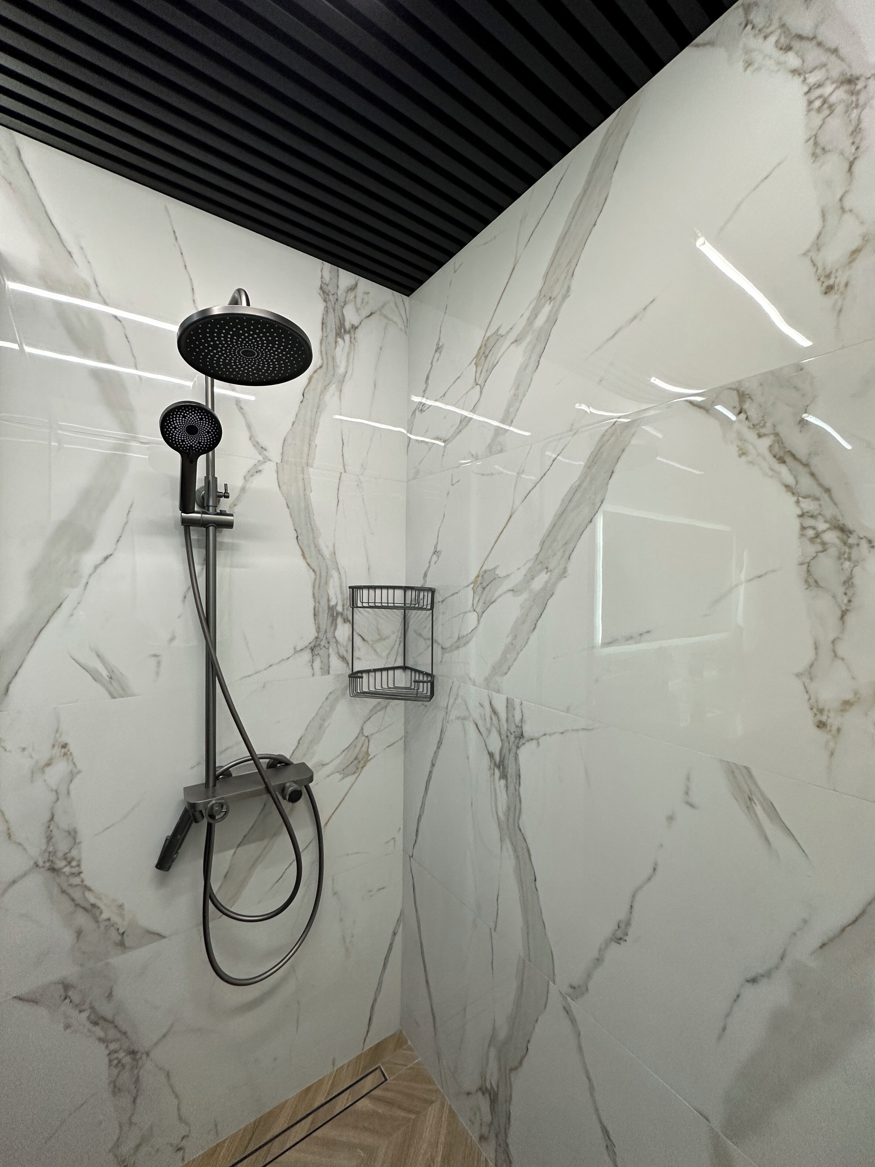 A modern bathroom with a marble wall, a shower with a handheld showerhead, and a wall-mounted shelf.