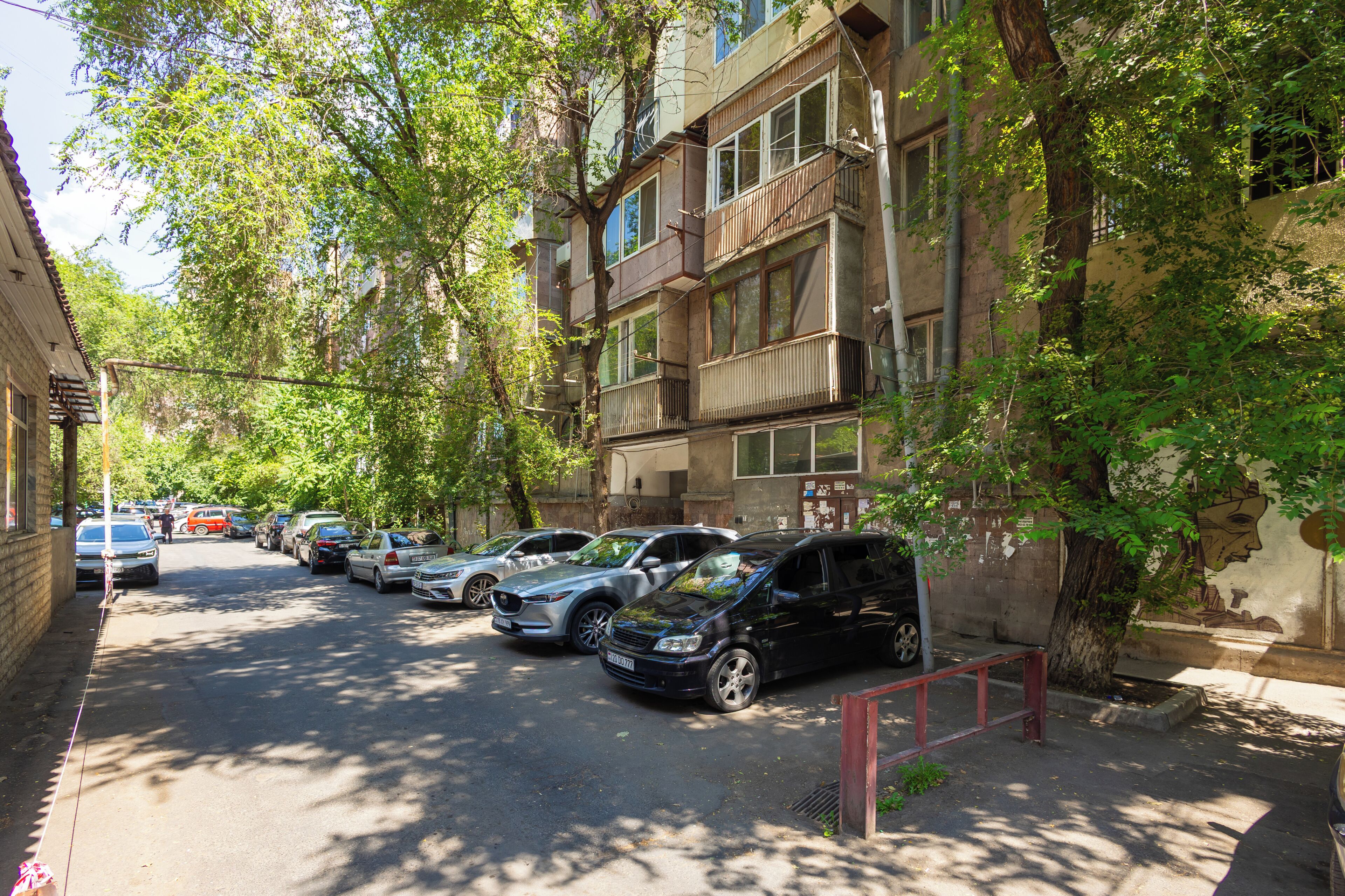 Une rangée de voitures garées le long d’une rue bordée d’arbres, à côté d’un immeuble à plusieurs étages doté de balcons.