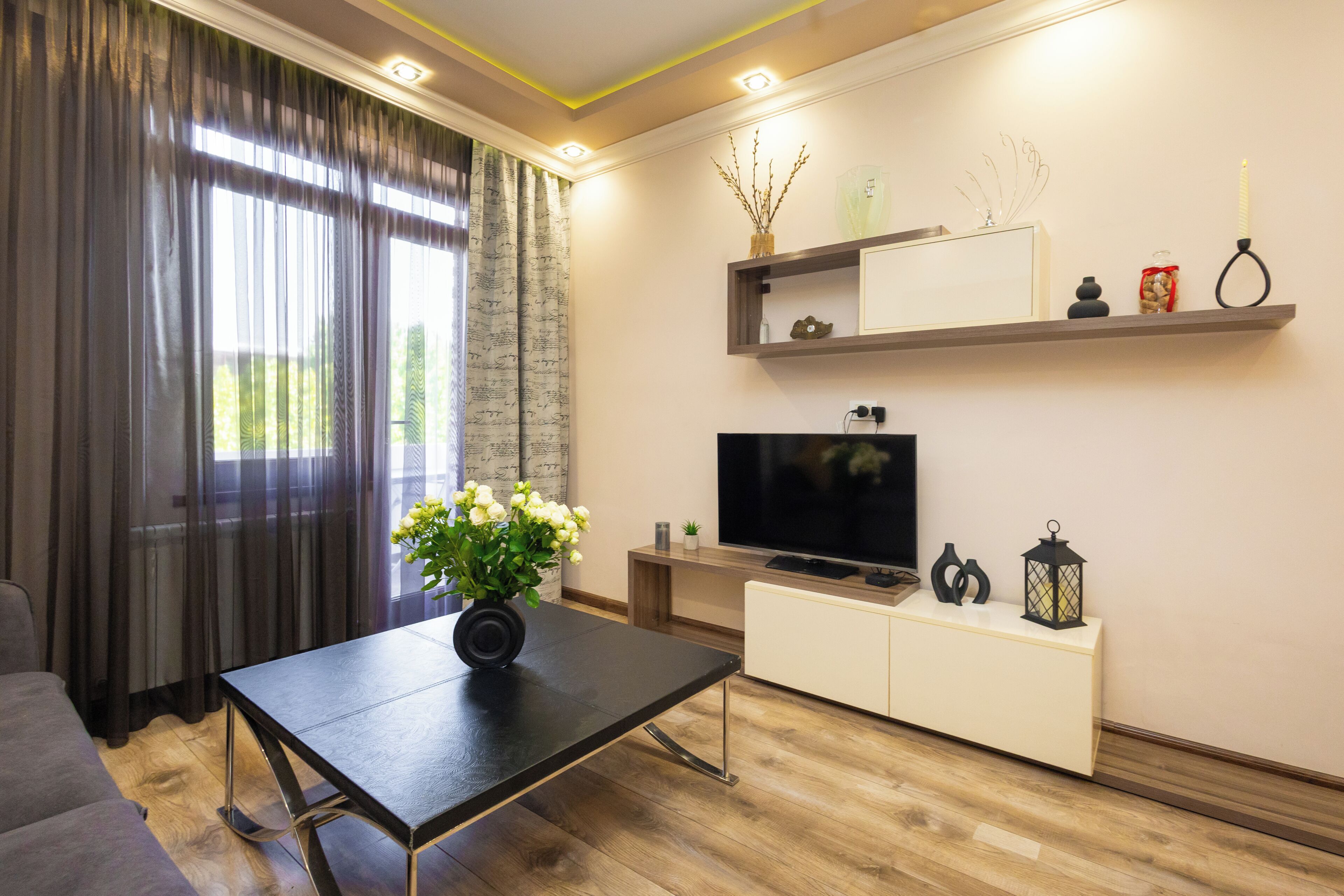 A modern living room with a flat-screen TV, a coffee table with a vase of flowers, a sofa, and a dining table.