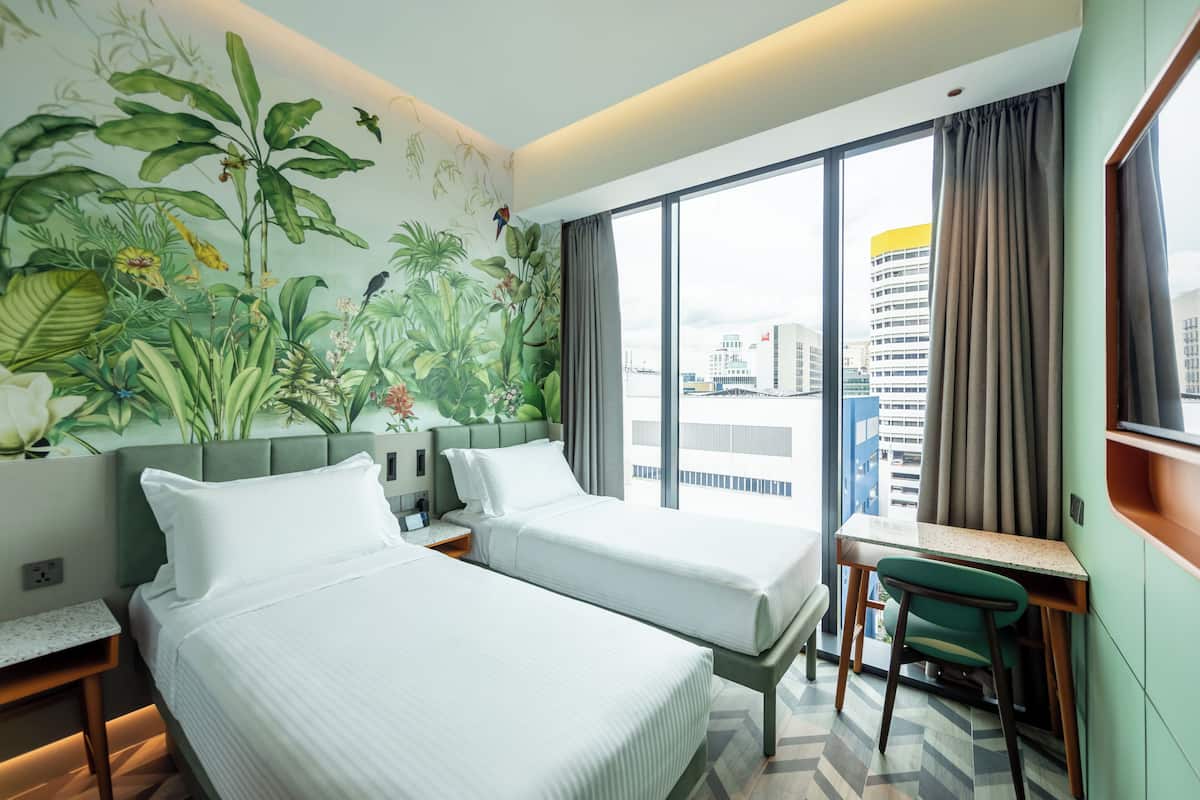 Une chambre d’hôtel avec deux lits, un bureau et une chaise. Il y a une grande fenêtre avec des rideaux, et une fresque représentant des plantes tropicales sur le mur.