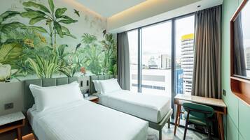 Quarto de hotel com duas camas, uma escrivaninha e uma cadeira. Há uma janela grande com cortinas, e um mural com plantas tropicais na parede.