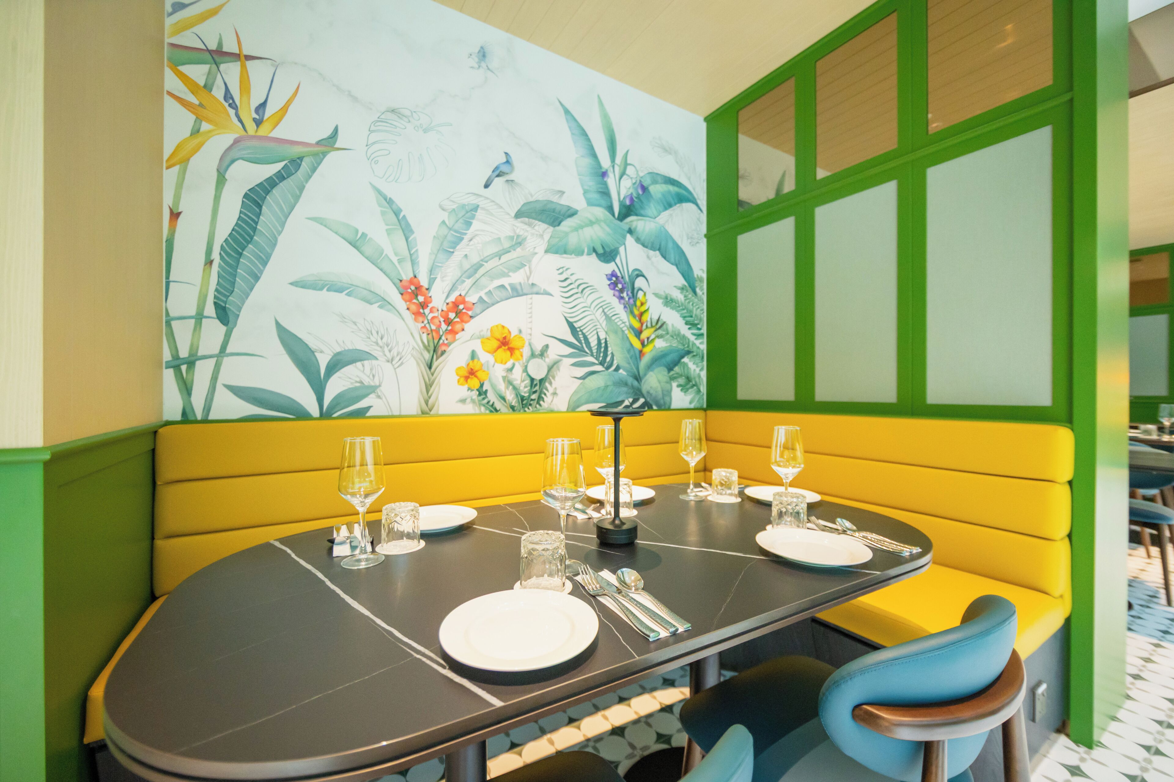 A dining area with a table set for four, green and yellow seating, and a mural with tropical plants.