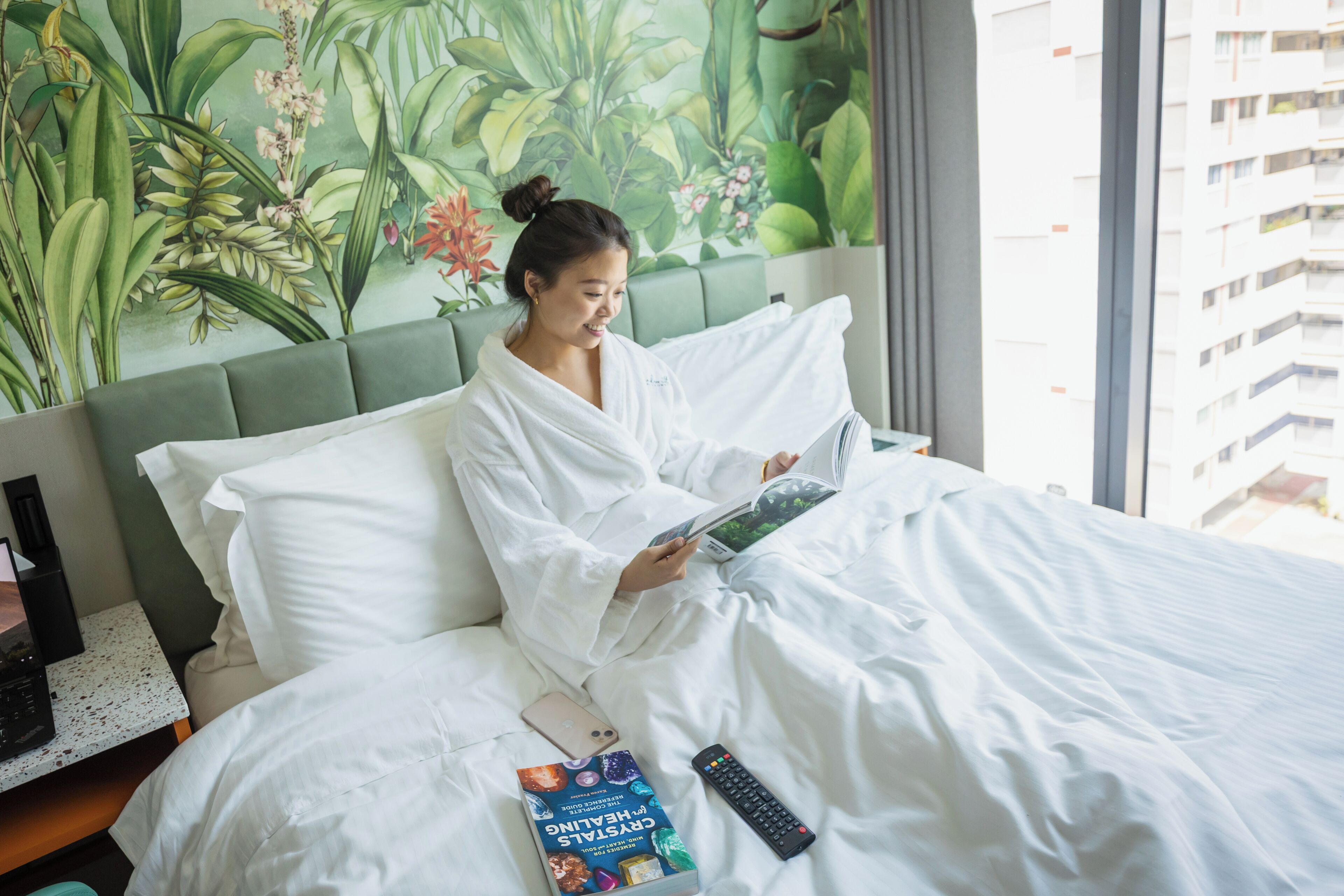 A person in a white bathrobe sitting on a bed with a book and a remote control, in a room with a green headboard and a floral wall mural.