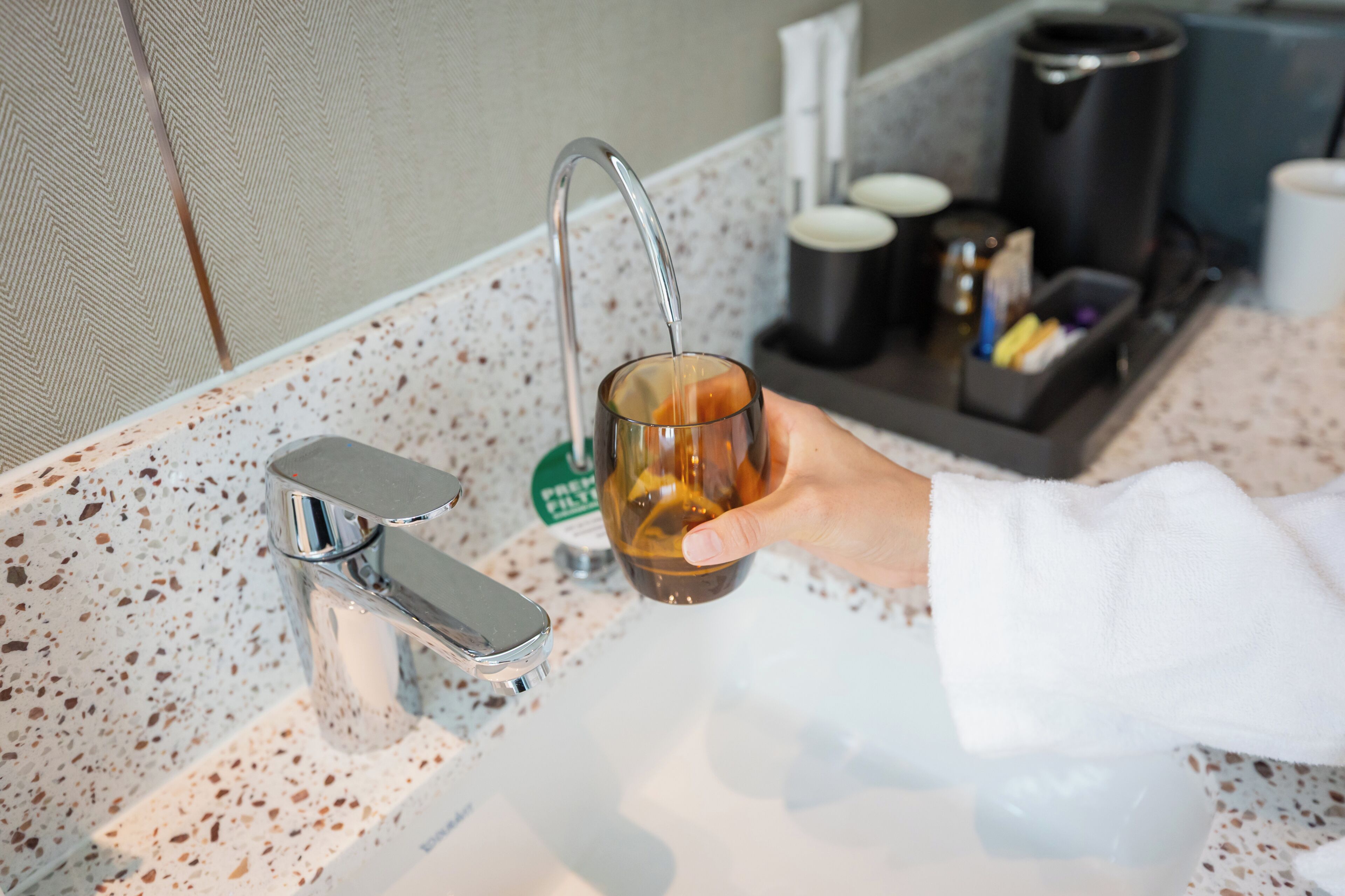 A hand holding a glass of amber liquid at a modern kitchen sink with a speckled countertop, a coffee maker, and a coffee pod holder.
