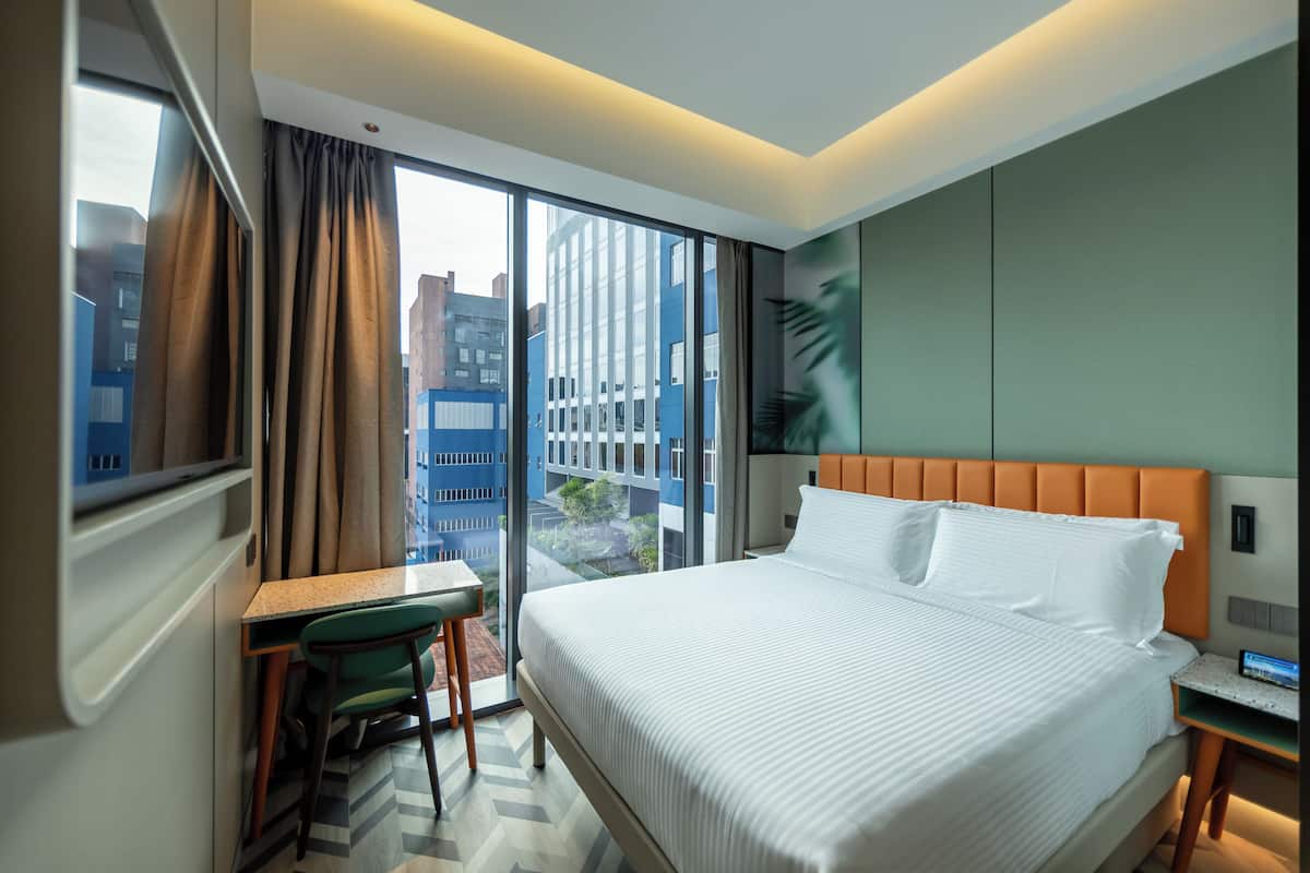 Une chambre d’hôtel moderne dotée d’un grand lit, d’un bureau et offrant une vue sur le paysage urbain grâce à de grandes fenêtres du sol au plafond.