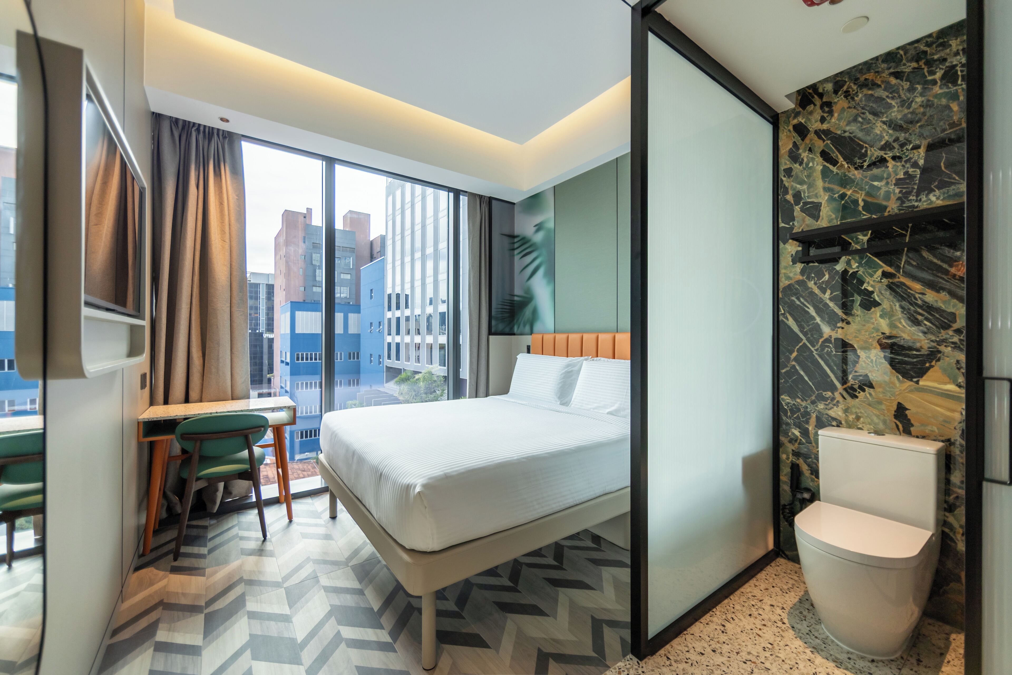 A modern hotel room with a bed, a desk with a chair, a toilet, and a large window offering a city view.