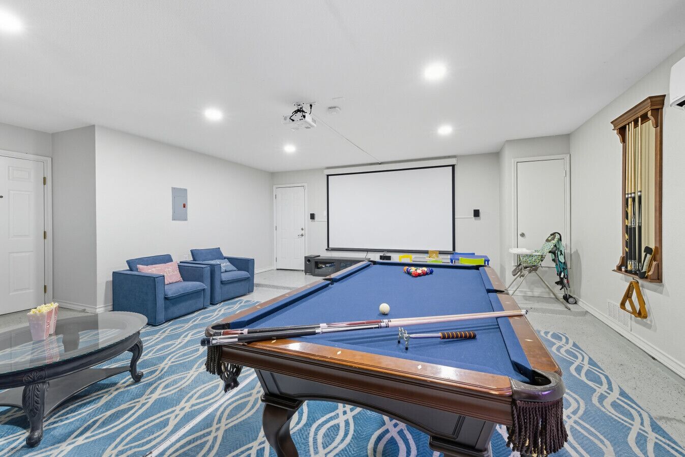 A pool table with a blue felt surface, a billiards cue, and a white cue ball. There is a projector screen on the wall and a pool table with a blue felt surface.
