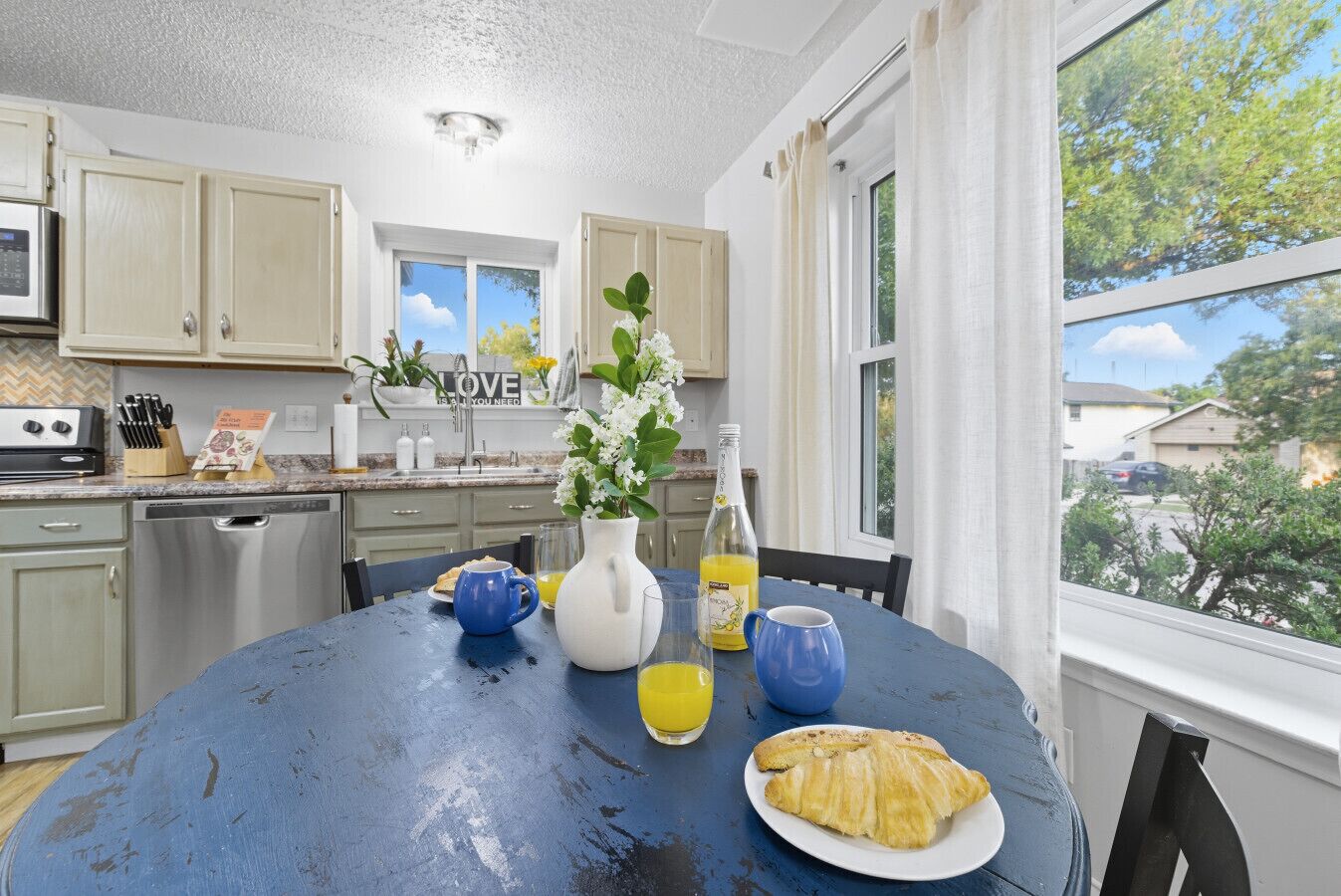 A kitchen with a blue table, a white pitcher, a bottle of orange juice, a blue mug, and a plate with a croissant.