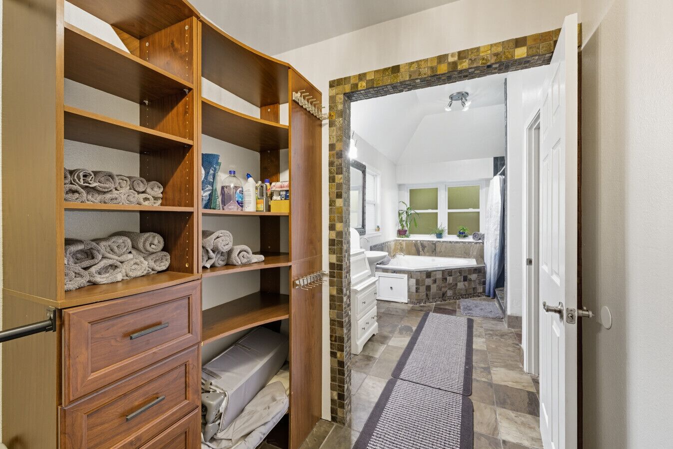 Un pasillo que conduce a un baño con bañera, lavabo y ventana. Se observa un mueble de madera con toallas y artículos de aseo.