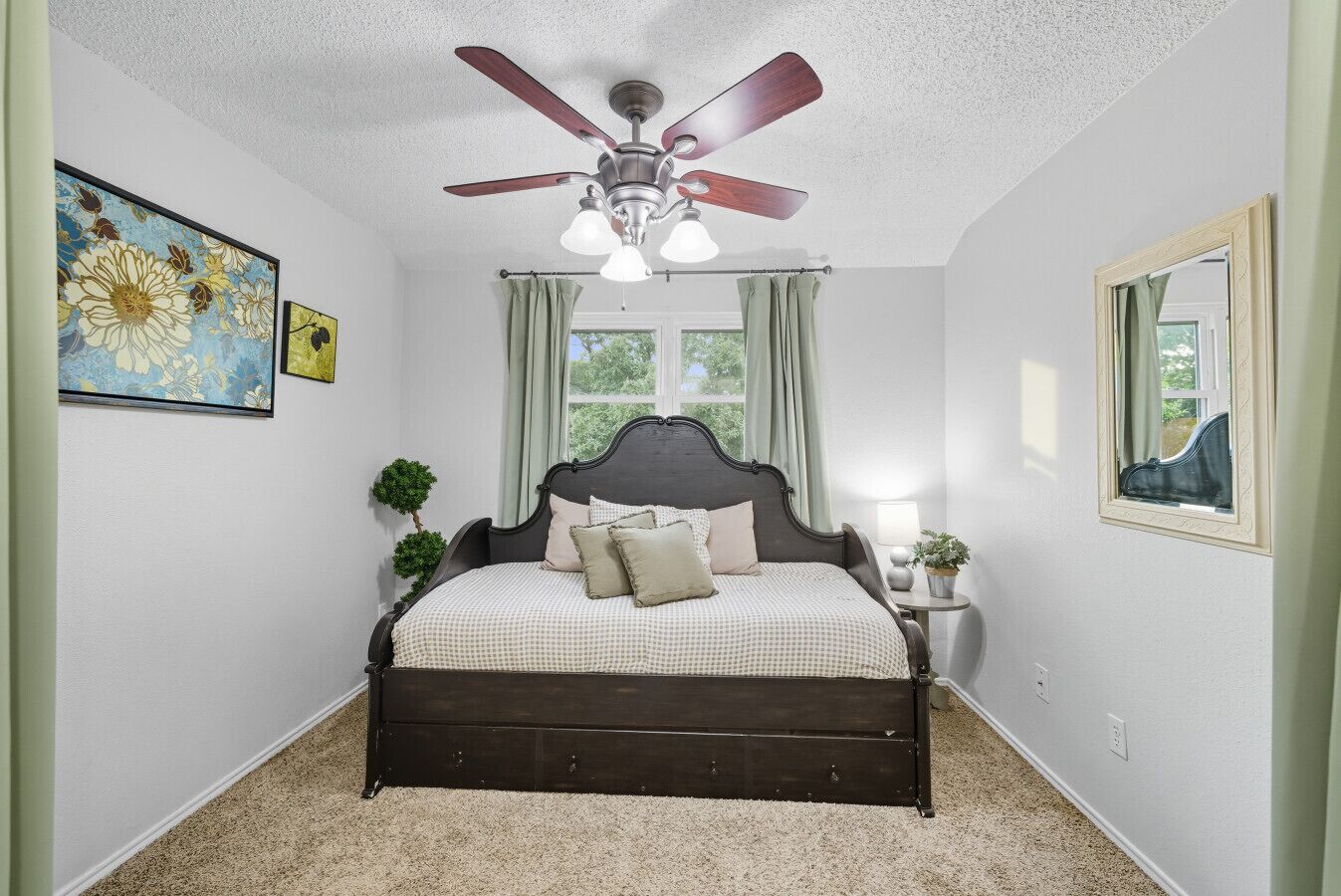 A bedroom with a dark wooden bed, a ceiling fan, a window with curtains, and a small bedside table with a lamp and a plant.