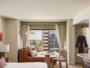 Habitación de hotel con cama, sofá, mesa de comedor, silla, lámpara, vista de un paisaje urbano y una bata colgada en la pared.