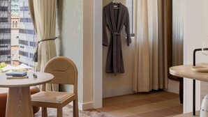 Une chambre d’hôtel avec vue sur les immeubles, une table ronde avec un livre, une chaise, un peignoir accroché au mur et un verre d’eau sur une table.