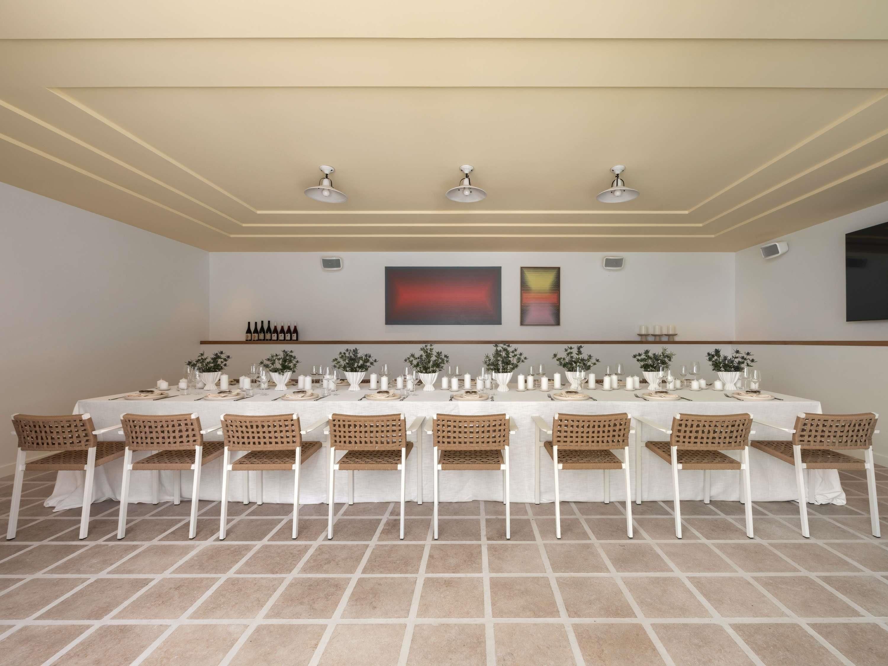 Una larga mesa de comedor con manteles blancos, rodeada de sillas de mimbre, en una habitación con piso de baldosas e iluminación empotrada en el techo.