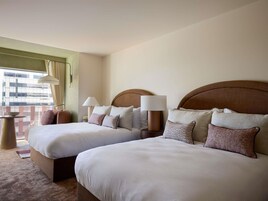 A hotel room with two beds, a window with curtains, a lamp, and a view of buildings.