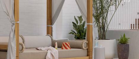 A wooden canopy bed with a beige mattress, a white curtain, and a pair of slippers on the floor.