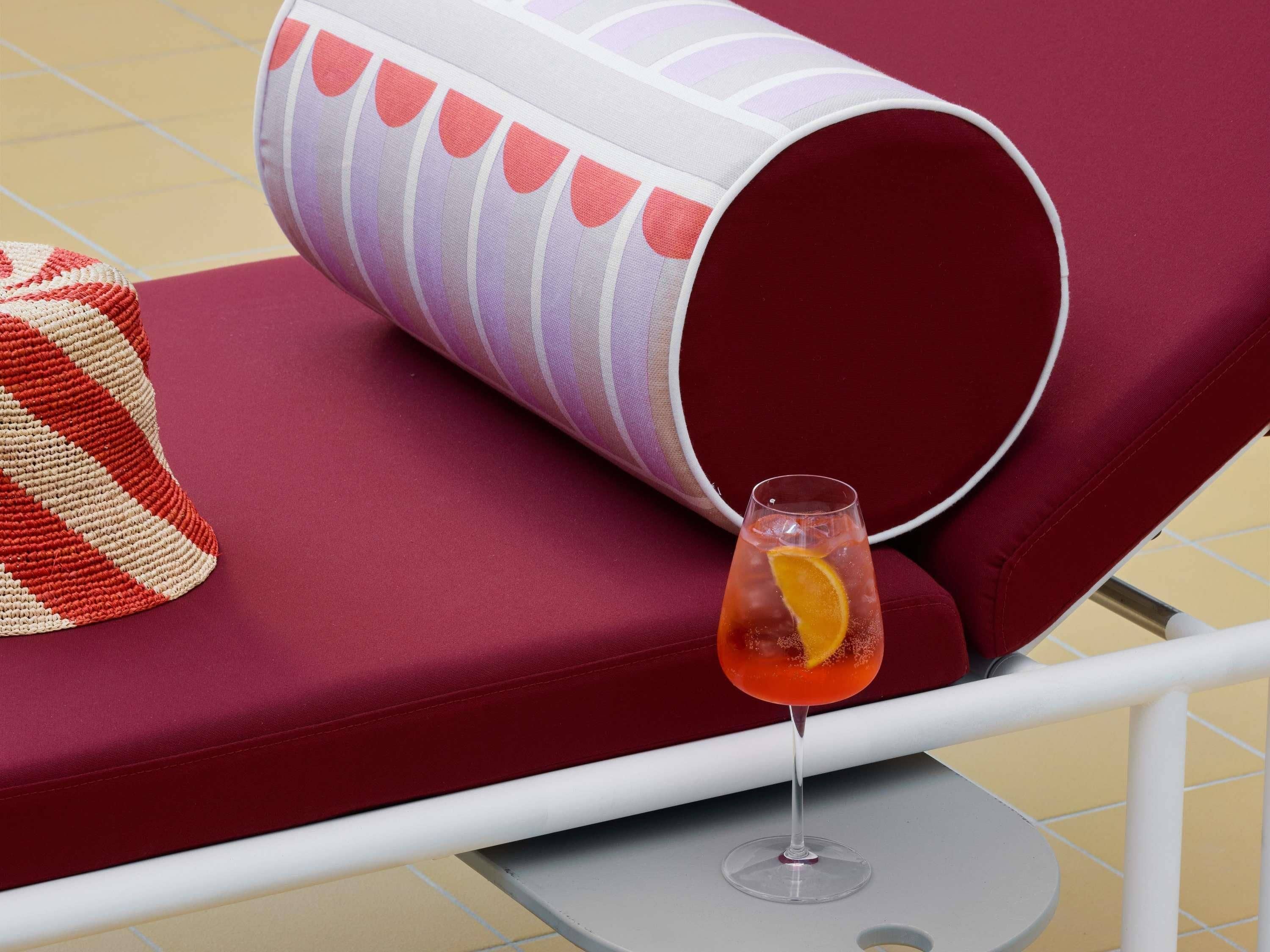 A lounge chair with a cushion, a glass of drink with an orange slice, and a striped towel.