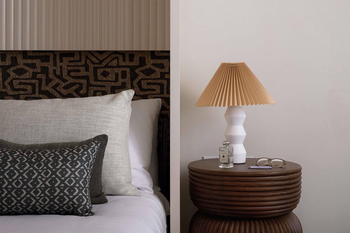 Un lit bien fait, agrémenté d’oreillers à motifs, une lampe moderne et une petite table avec des verres et une bouteille.