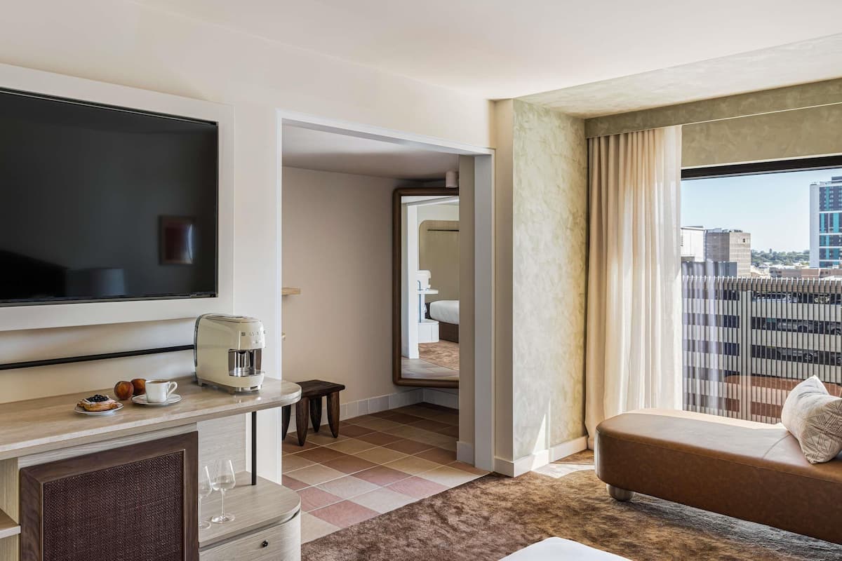 A hotel room with a flat-screen TV, a coffee maker, a dining area, a sofa, and a city view through the window.