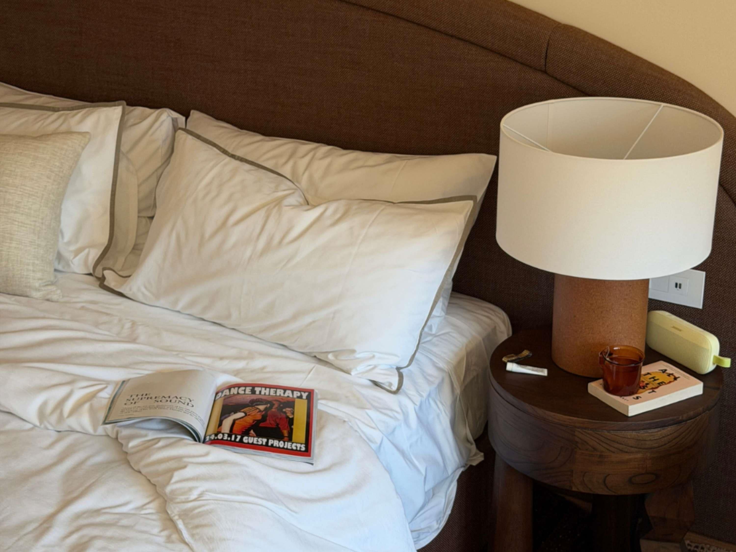 A neatly made bed with white linens, a magazine on the bed, a lamp on a wooden nightstand, and a book beside the lamp.