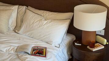 A neatly made bed with white linens, a magazine on the bed, a lamp on a wooden nightstand, and a book beside the lamp.