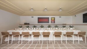 Een lange eettafel met witte tafelkleden, omringd door rieten stoelen, in een kamer met een betegelde vloer en een plafond met inbouwverlichting.